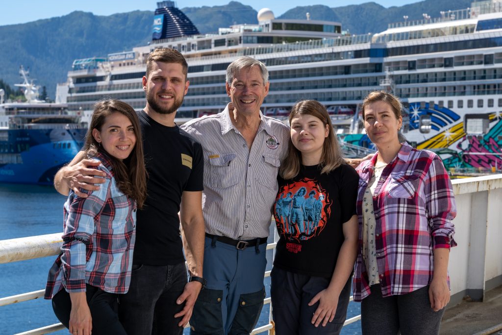 Five people - four Ukrainian refugees and an Alaska businessman - stand on the deck of a ferry as a cruise ship provides the background